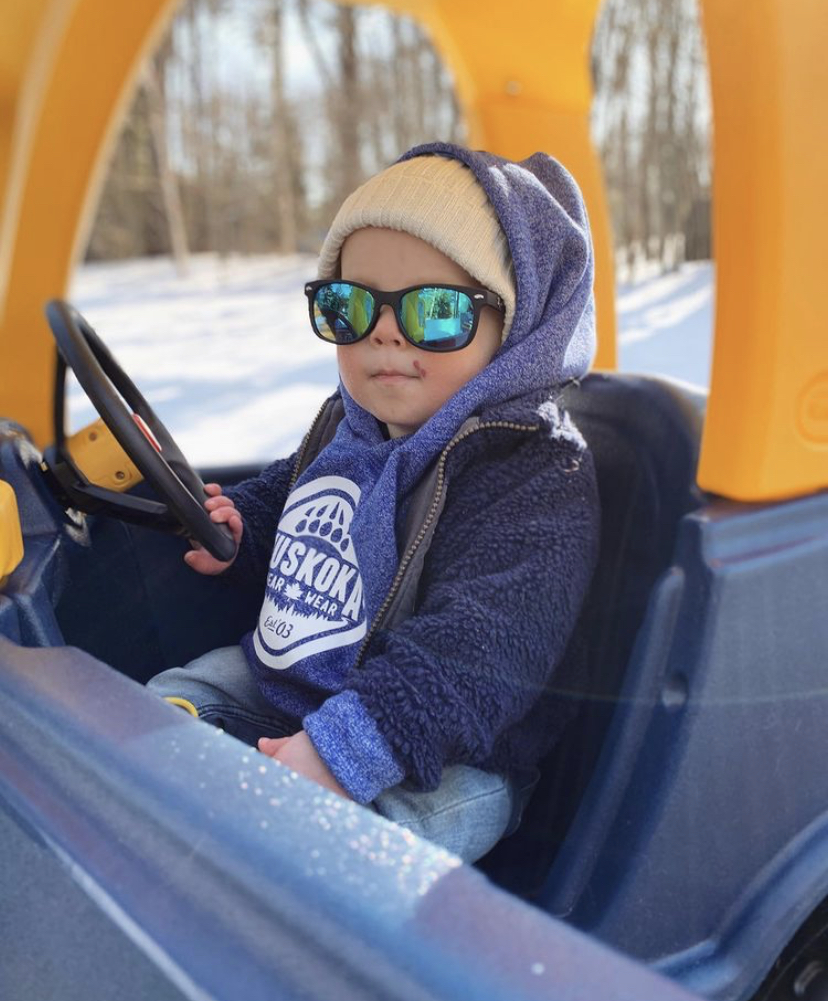 Toddler wearing a Muskoka Bear Wear hoody and some sunglasses while driving a play car
