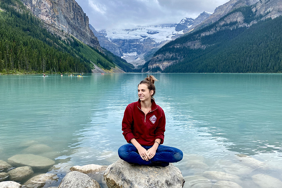 Canadian woman sitting on a rock wearing jeans and a red quarter zip sweater from Muskoka Bear wear. She is sitting in front of a lake with trees and mountain in the background