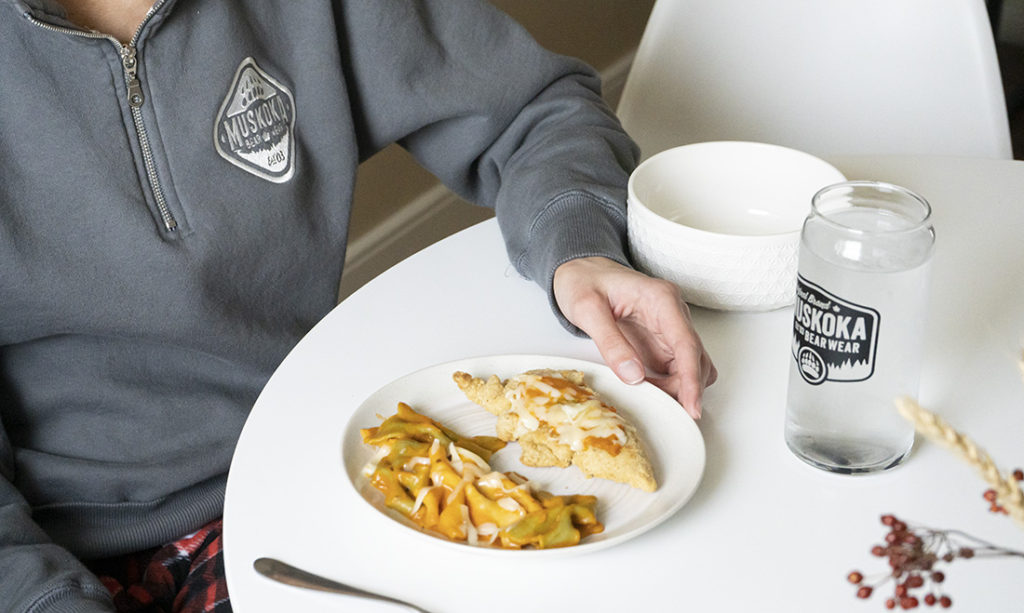 Close-up of person sitting at a dinner table with chicken Parmesan and pasta, a MBW glass full of water, wearing a dark grey ladies quarter-zip and red cottage comfy pants.